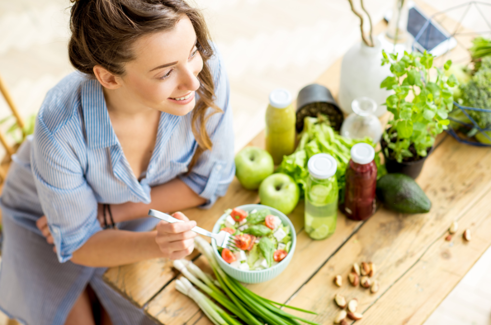 A woman sitting at a wooden table with a variety of fruits and vegetables sitting beside her salad.