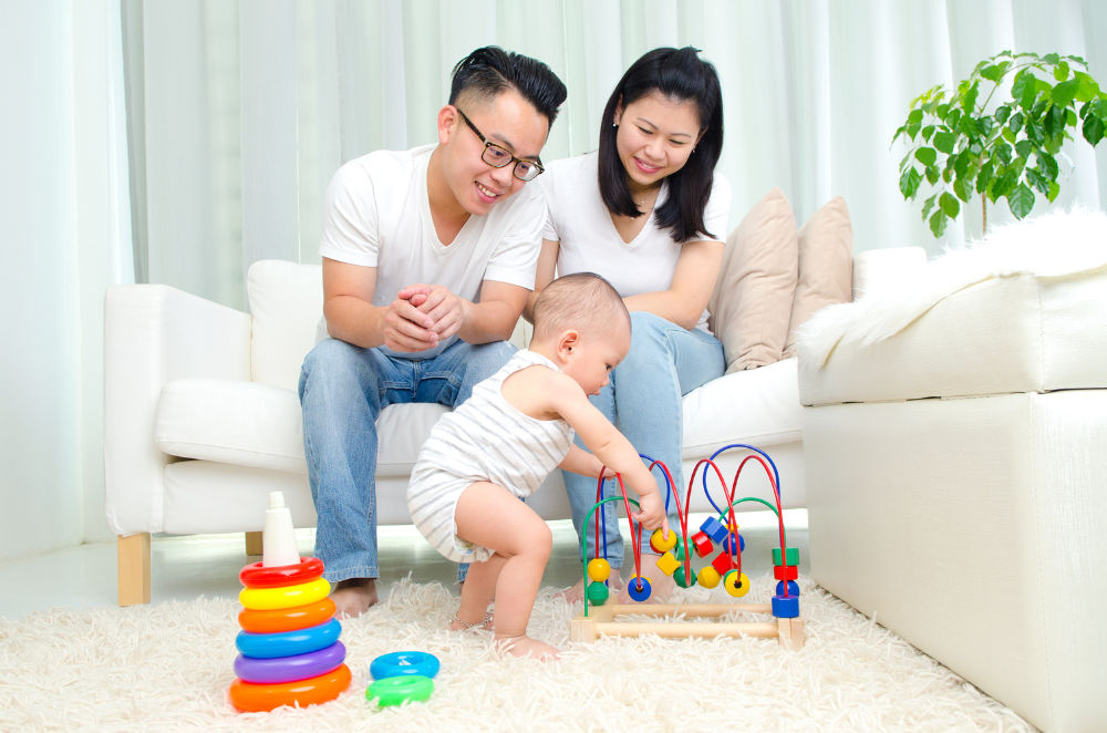 Parents sitting on a couch looking at their baby playing on the ground in their living room.
