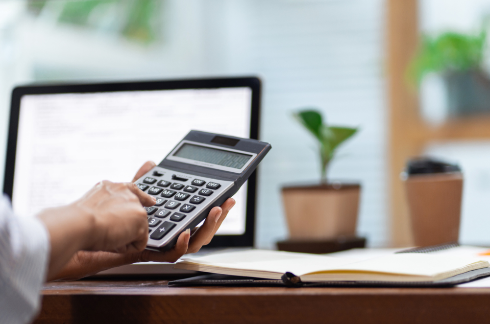 A hand typing on a calculator while the person sits at a desk with a laptop and plant in the background on the desk.