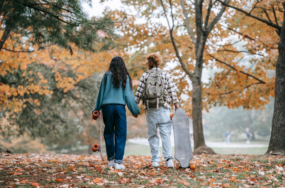 A couple walking in fall leaves with longboards in their hands.